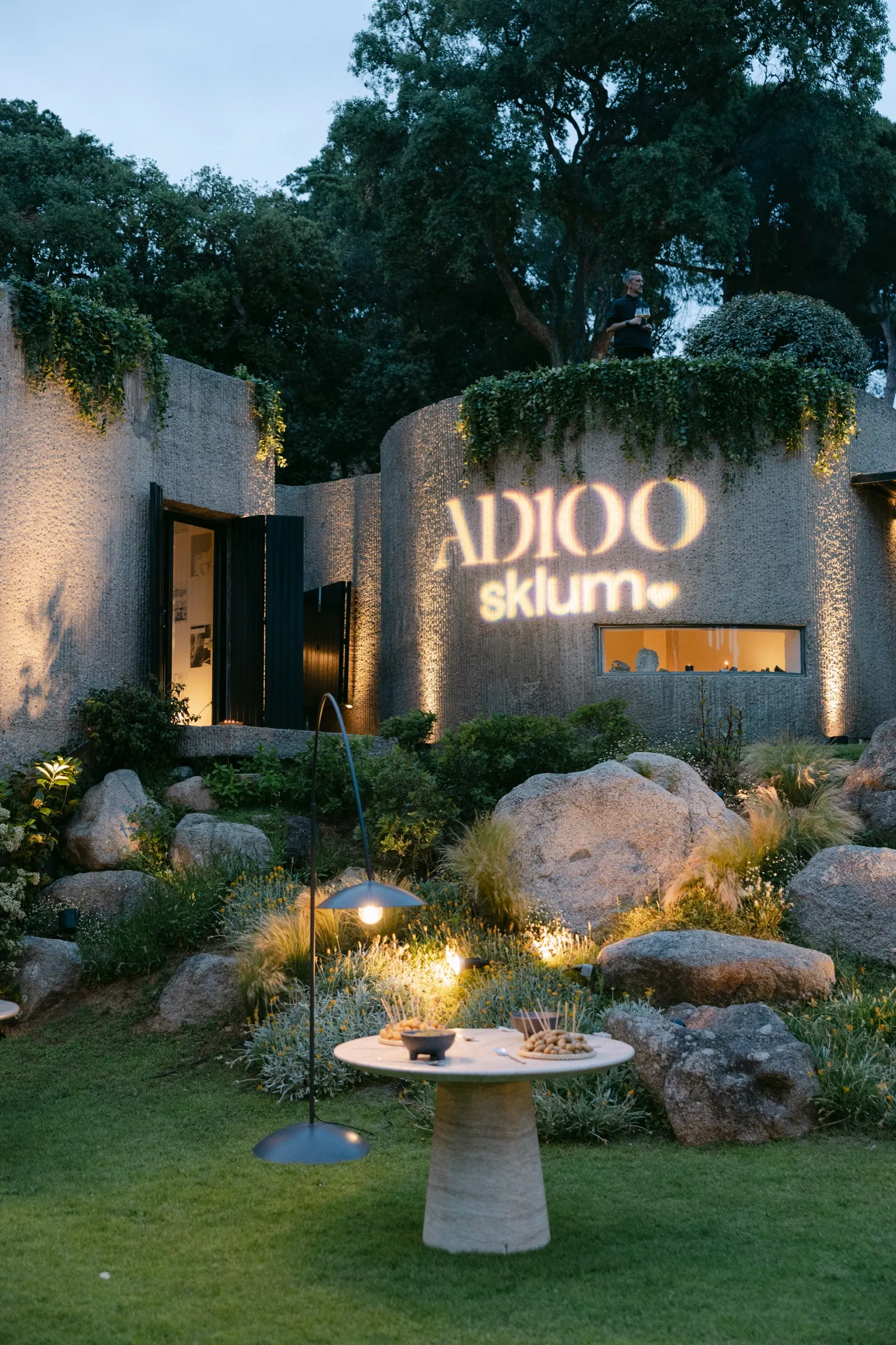 Rótulo luminoso de AD100 y Sklum en la fachada de la Casa Semienterrada al anochecer. Fotografía de Noomadik.