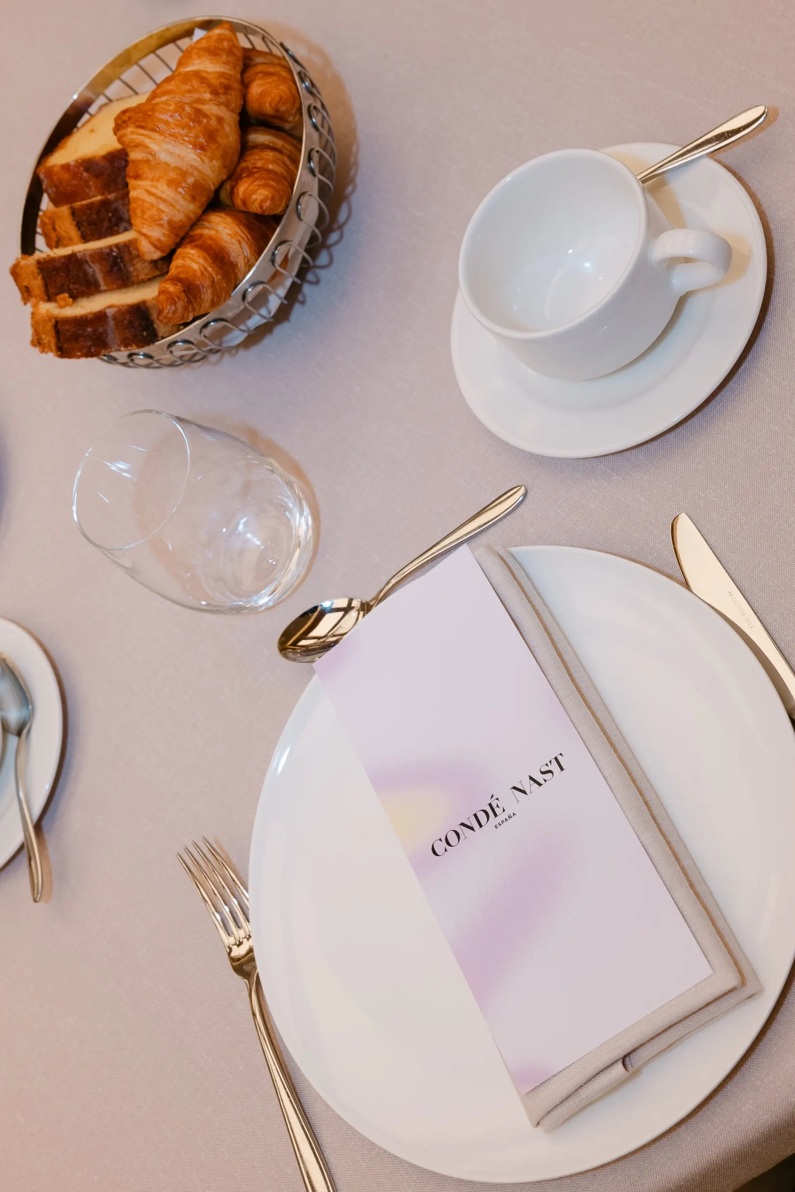 Vista cenital de cruasanes, taza y menú de Condé Nast sobre la mesa del brunch. Fotografía de Noomadik.
