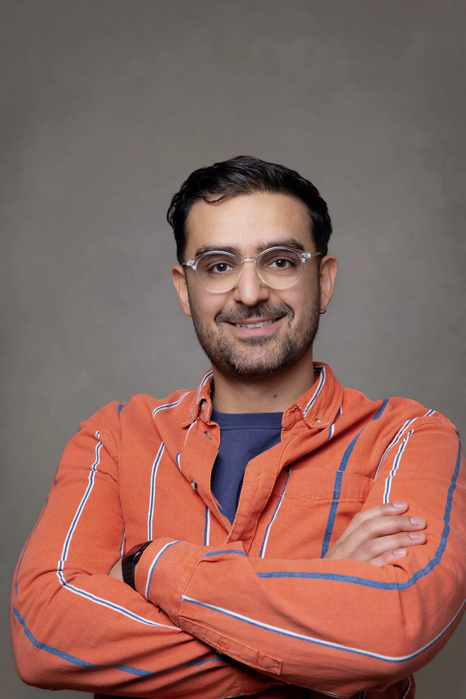Retrato corporativo de un hombre con gafas y camisa naranja de brazos cruzados sobre fondo gris. Fotografía de Noomadik.